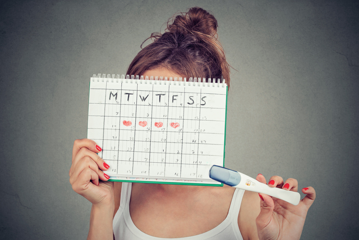 Young woman hiding behind a periods calendar and showing a positive pregnancy test, successfully knowing how to tell when you're ovulating