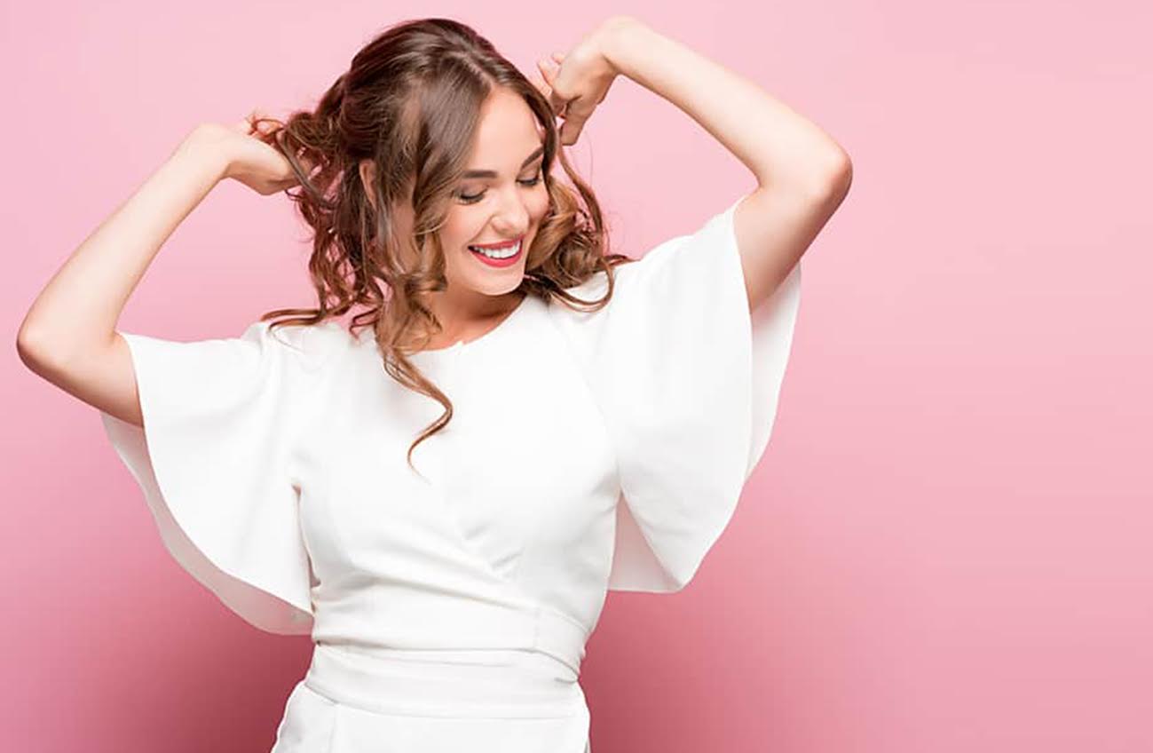 A happy woman holding up her hair, wearing white on a a pink background, learning best practices to keep your vagina clean.