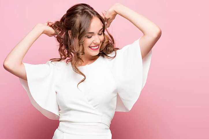 A happy woman holding up her hair, wearing white on a a pink background, learning best practices to keep your vagina clean.