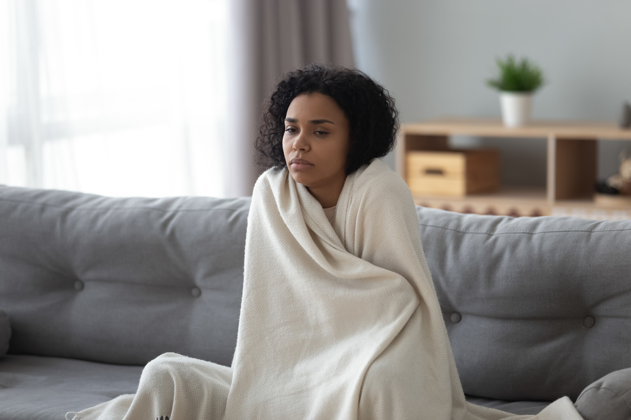 African American woman sitting on couch wrapped up in blanket, experiencing winter blues.