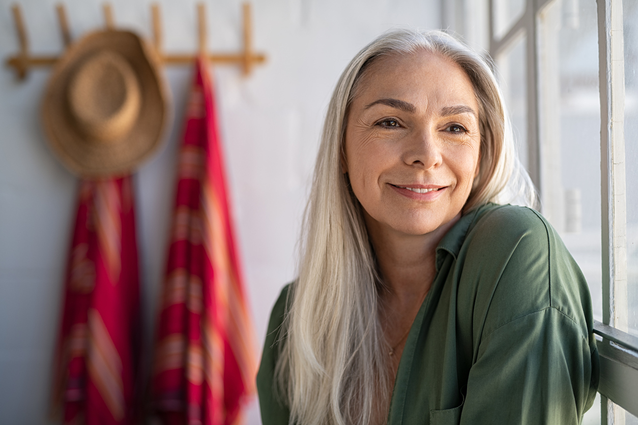 Portrait of beautiful senior woman sitting near big window at home and contemplate. Closeup face of fashionable old woman at home relaxing in living room. Mature woman thinking while looking away, wondering what the Most Common Health Issues Women Face in their 50s are.