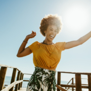 Excited young woman running on a boardwalk with her hands raised on a sunny day. African female having fun on summer vacation at the seaside, knowing how your happiness can impact your health.