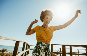 Excited young woman running on a boardwalk with her hands raised on a sunny day. African female having fun on summer vacation at the seaside, knowing how your happiness can impact your health.