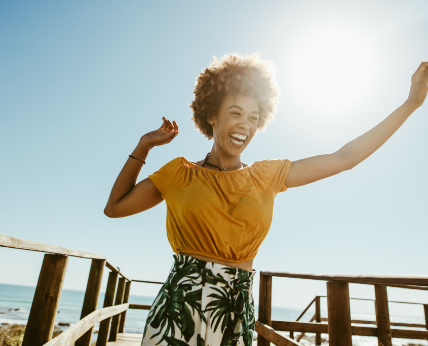 Excited young woman running on a boardwalk with her hands raised on a sunny day. African female having fun on summer vacation at the seaside, knowing how your happiness can impact your health.