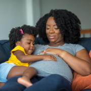 African American mother and daughter smiling at home on the couch, both with their hand on the woman's pregnant belly, knowing the The Effects of Systemic Racism on Black Women’s Healthcare Access.