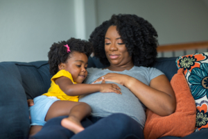 African American mother and daughter smiling at home on the couch, both with their hand on the woman's pregnant belly, knowing the The Effects of Systemic Racism on Black Women’s Healthcare Access.