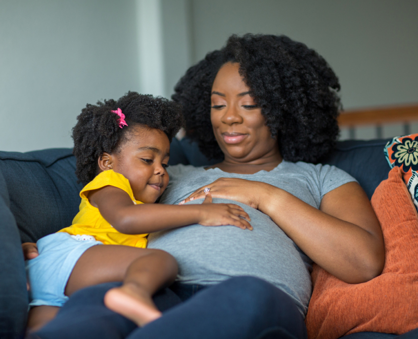 African American mother and daughter smiling at home on the couch, both with their hand on the woman's pregnant belly, knowing the The Effects of Systemic Racism on Black Women’s Healthcare Access.