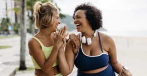 Two happy women in sportswear enjoy their workout routine on the ocean promenade, celebrating with a high-five. Female friends with different body shapes embracing a lifestyle of fitness.